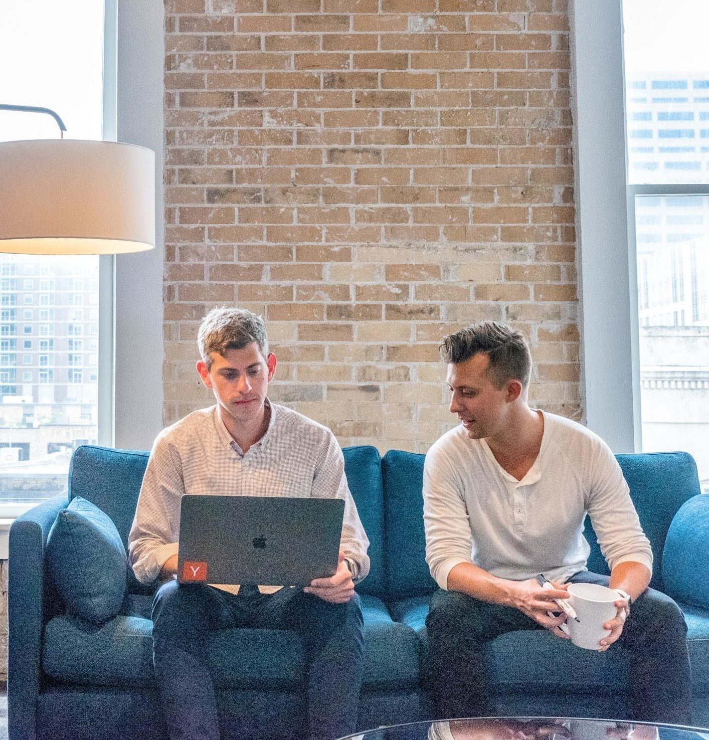 two men sitting on a blue sofa looking at a macbook