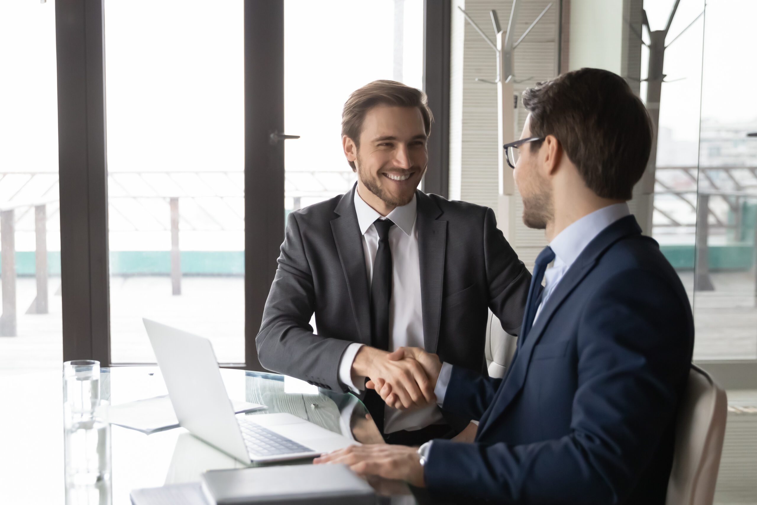 two men in suits sitting at table shaking hands and smiling