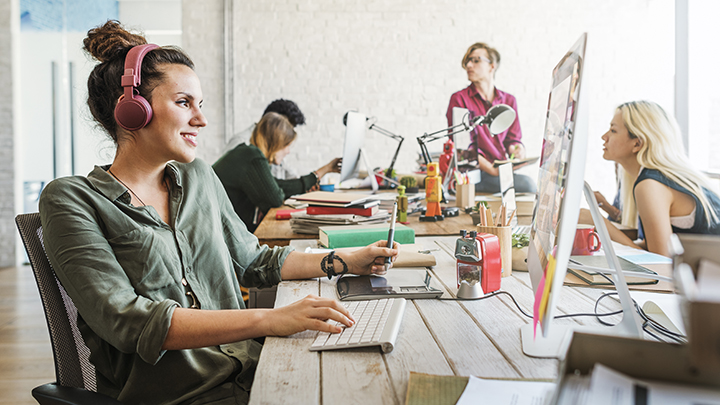 woman working on computer with headphones on among other coworkers